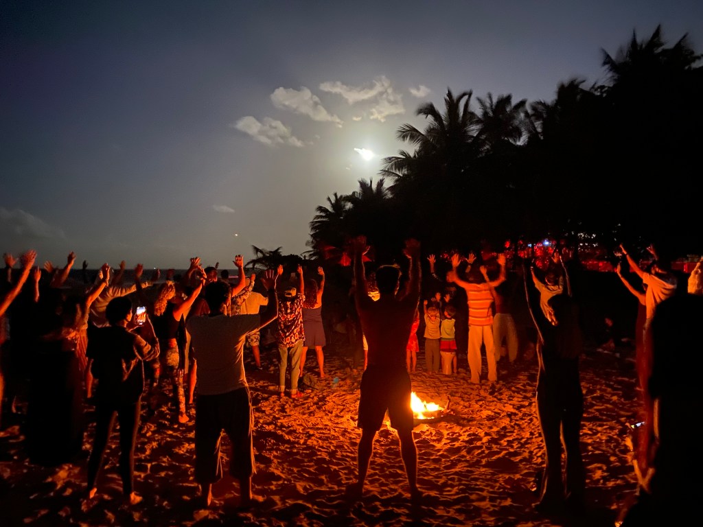A group of people gathered on a beach at night, raising their arms in celebration around a fire, with a full moon illuminating the scene.